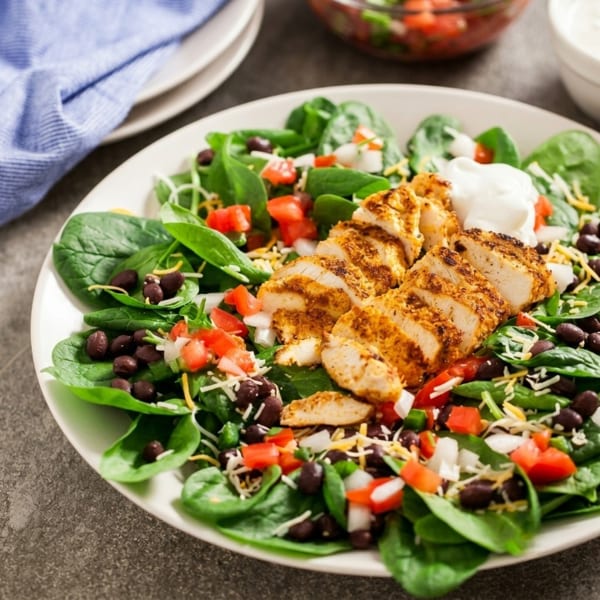 A vibrant salad with sliced seasoned chicken, spinach, black beans, tomatoes, cheese, and sour cream.