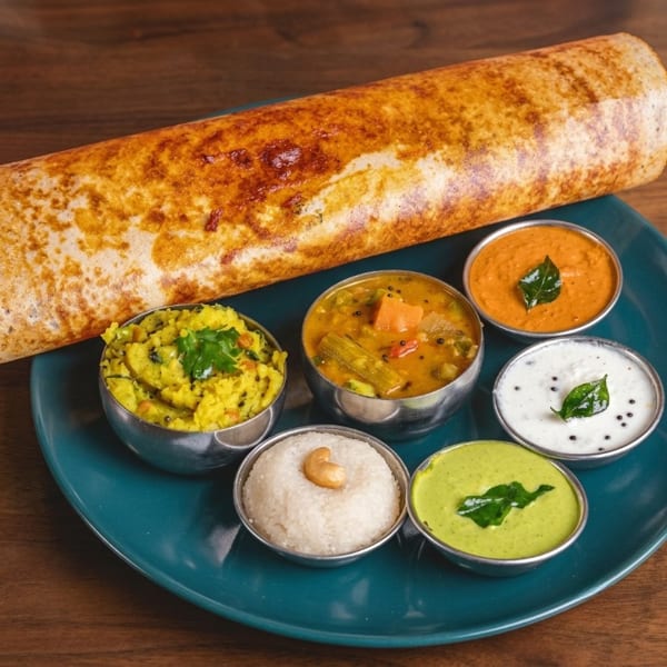 Golden-brown dosa served with six small bowls of colorful chutneys and sambar on a teal plate.
