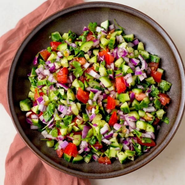 Freshly chopped salad with cucumbers, tomatoes, red onions, and herbs in a dark ceramic bowl.